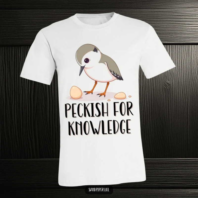Funny t-shirt depicting a sandpiper character curiously pecking at a small shell, highlighting a moment of gentle discovery.